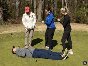 3 women golfers pointing clubs at male golfer on ground