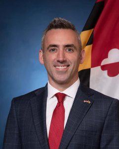 headshot of male wearing suit with maryland flag behind him