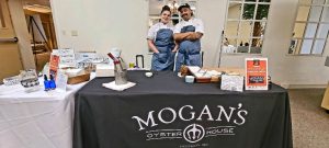 A man and woman standing in front of a table with the words Mogan's on the table cloth