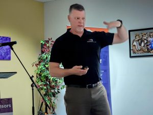 Man in a black shirt speaking at a luncheon