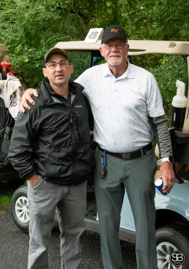 Man in a white polo shirt standing with a man in a black windbreaker jacket in front of a golf cart