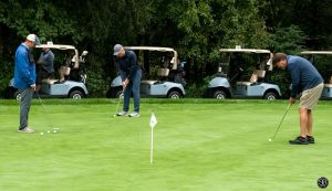 Three men putting golf balls on a golf course with golf carts in the background
