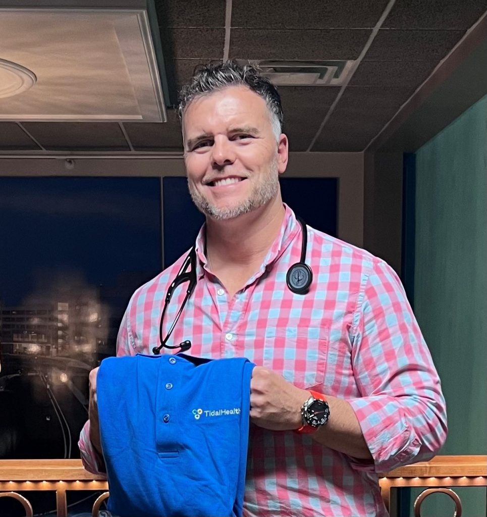 Headshot of Ryan Brown wearing a flannel shirt while holding a blue TidalHealth shirt