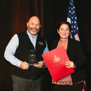 Man and Woman on stage beside an American flag holding an award