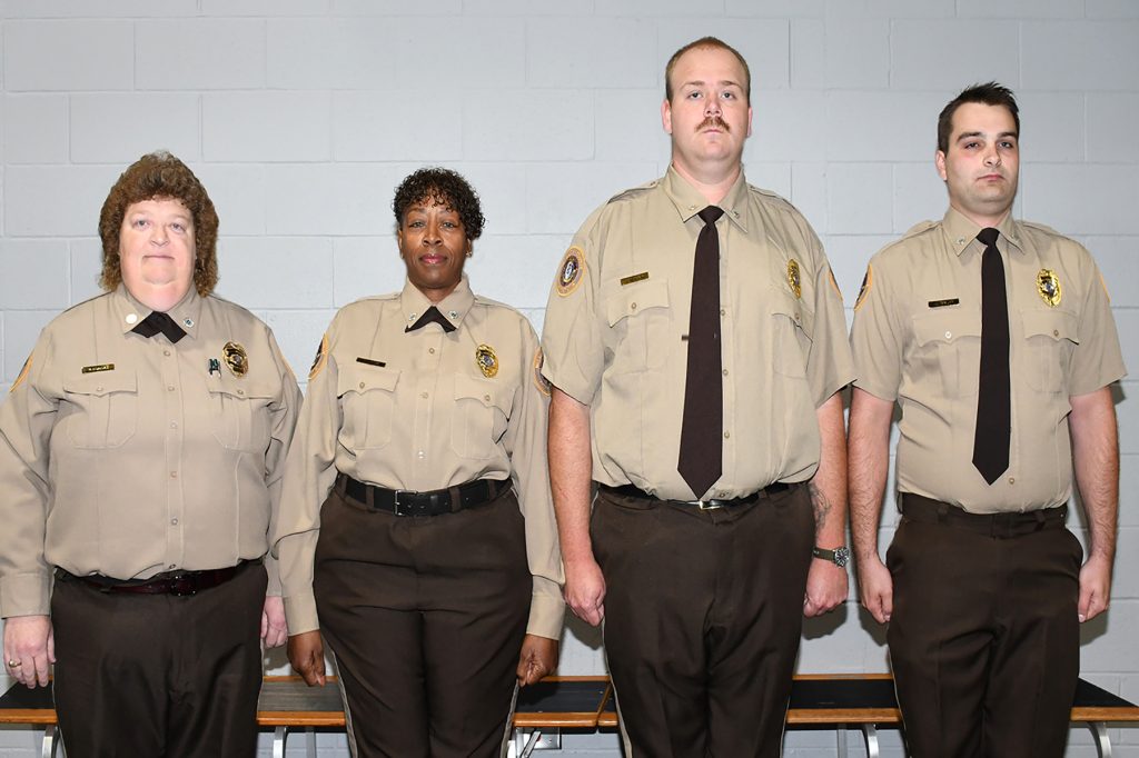 four correctional officers in uniform standing against a white wall