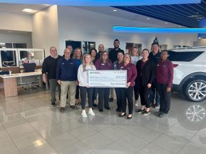 Large group of men and women in a car dealership holding a large check