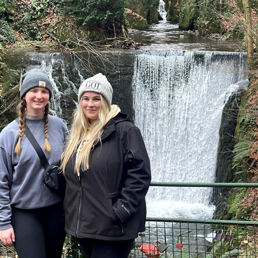 two female students standing outside beside a waterfall 