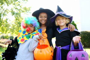 Three young children smiling outdoors in Halloween costumes: a clown with a colorful wig and large bowtie, and two witches holding pumpkin-shaped trick-or-treat buckets.