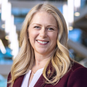 Headshot of Carolyn Ringer Lepre smiling, wearing a maroon blazer and a white top against a softly blurred background.