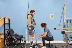 A physical therapist assists a young man using a walker and an overhead safety harness in a therapy clinic equipped with a wheelchair and exercise gear.