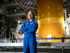 NASA astronaut Christina Koch standing with arms crossed in a bright blue NASA flight suit, looking confident and smiling off to the side. Behind her is a massive, cylindrical orange Space Launch System (SLS) rocket core stage inside a large industrial assembly facility with steel scaffolding and metal railings.