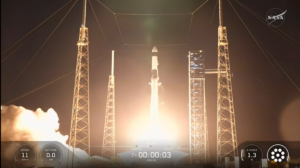 NASA webcast screenshot showing the night launch of a SpaceX Falcon 9 rocket for the CRS-35 commercial resupply mission. A bright, glowing plume of fire and thick smoke illuminates the launchpad as the rocket lifts off between tall metal lightning towers and a service structure. A mission telemetry overlay at the bottom displays "T+ 00:00:03", speed, altitude, and G-force stats, with a NASA logo in the top right corner.