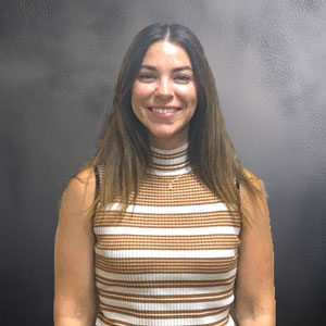 sarah-brissey-crm Headshot of Sarah Brissey smiling directly at the camera with long brown hair, wearing a sleeveless high-neck ribbed top with brown and white horizontal stripes against a textured dark gray background.
